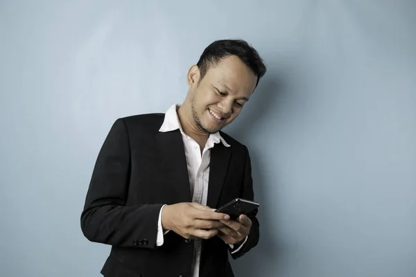 A portrait of a happy Asian businessman is smiling and holding his smartphone wearing black suit isolated by a blue background