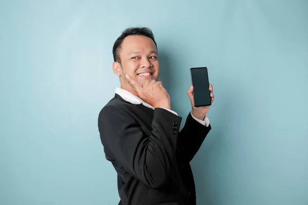 A portrait of a happy Asian businessman is smiling and holding his smartphone pointing copy space beside him wearing black suit isolated by a blue background