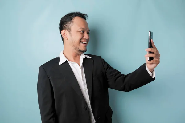 A portrait of a happy Asian businessman is smiling and holding his smartphone wearing black suit isolated by a blue background