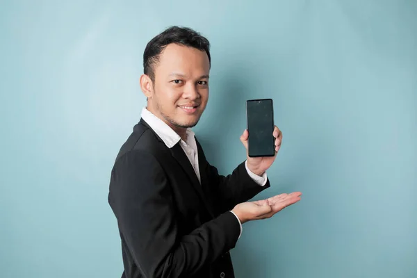 A portrait of a happy Asian businessman is smiling and holding his smartphone showing copy space on it\'s screen wearing black suit isolated by a blue background