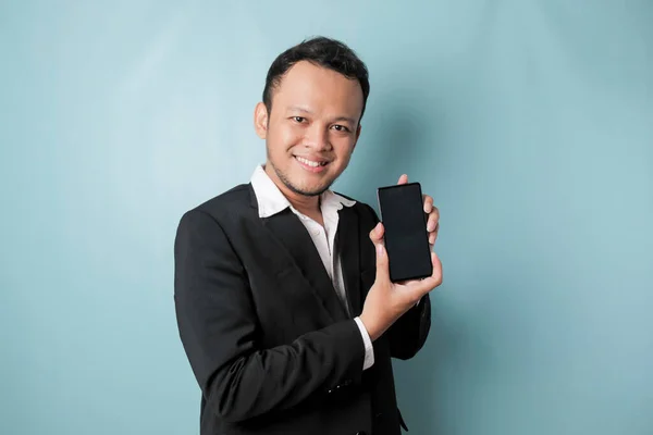 A portrait of a happy Asian businessman is smiling and holding his smartphone showing copy space on it\'s screen wearing black suit isolated by a blue background