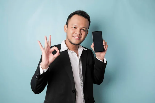 A portrait of a happy Asian businessman is smiling and holding his smartphone showing copy space on it\'s screen and gesturing Ok sign wearing black suit isolated by a blue background