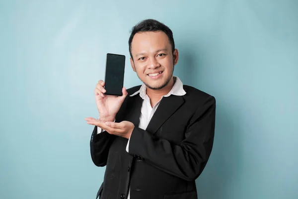 A portrait of a happy Asian businessman is smiling and holding his smartphone showing copy space on it\'s screen wearing black suit isolated by a blue background