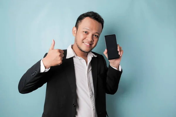 A portrait of a happy Asian businessman is smiling and holding his smartphone showing copy space on it's screen wearing black suit isolated by a blue background