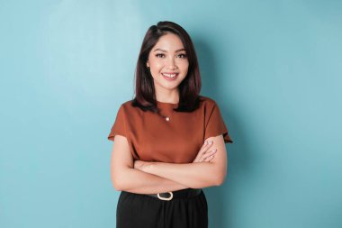 Portrait of a confident smiling Asian woman standing with arms folded and looking at the camera isolated over blue background