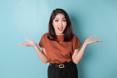 A portrait of an Asian woman wearing a brown shirt looks so confused, isolated by a blue background