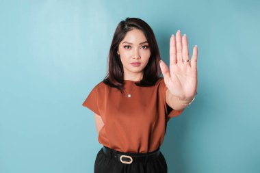 Young Asian woman wearing brown shirt over blue isolated background doing stop sign with palm of the hand.