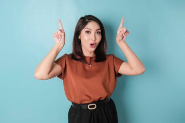 Shocked Asian woman wearing pointing at the copy space upside her, isolated by blue background
