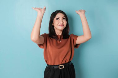 Excited Asian woman wearing brown shirt pointing at the copy space upside her, isolated by blue background