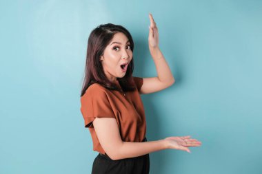 Shocked Asian woman pointing at the copy space beside her and smiling, isolated by blue background