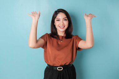 Excited Asian woman wearing brown shirt pointing at the copy space upside her, isolated by blue background