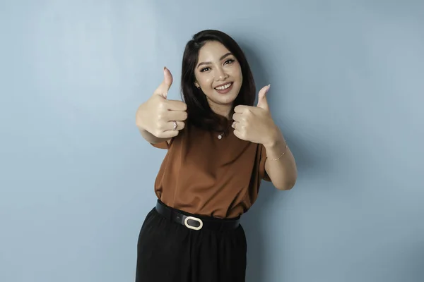Excited Asian woman gives thumbs up hand gesture of approval, isolated by blue background