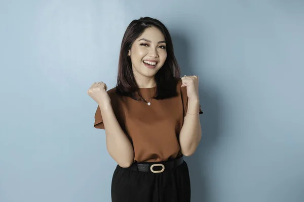 A young Asian woman with a happy successful expression wearing brown shirt isolated by blue background