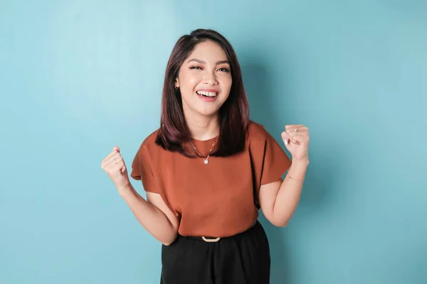 A young Asian woman with a happy successful expression wearing brown shirt isolated by blue background