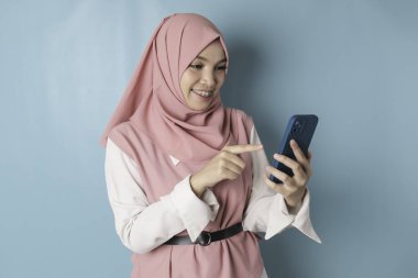 A young Asian Muslim woman wearing pink hijab is smiling while holding her smartphone