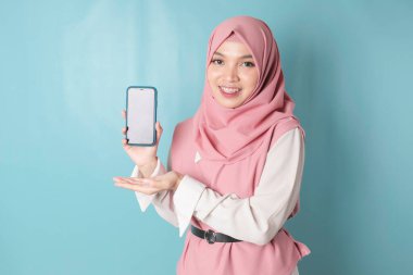 A young Asian Muslim woman wearing pink hijab is smiling while showing her smartphone screen