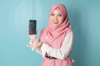 A young Asian Muslim woman wearing pink hijab is smiling while showing her smartphone screen