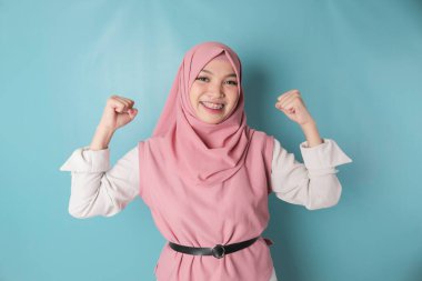 Excited Asian Musllim woman wearing a pink hijab showing strong gesture by lifting her arms and muscles smiling proudly