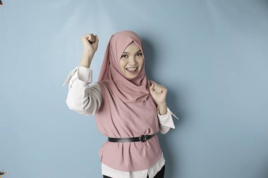 A young Asian Muslim woman with a happy successful expression wearing pink hijab isolated by blue background