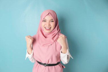 A young Asian Muslim woman with a happy successful expression wearing pink hijab isolated by blue background