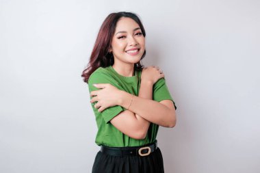 Genç, güzel Asyalı bir kadın, beyaz arka plan üzerine günlük yeşil tişört giyip mutlu ve pozitif bir şekilde kendine sarılıyor, kendinden emin gülümsüyor. Sevgi ve özgüven.