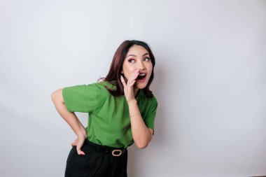 Yeşil bir tişört giyen genç ve güzel bir kadın bağırıyor ve bir eli ağzında bağırıyor. iletişim konsepti.