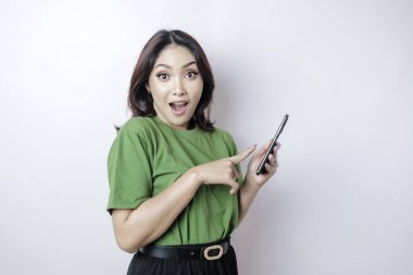 Yeşil tişört giyen Asyalı kadın akıllı telefonunu işaret ediyor, beyaz arka planla izole edilmiş.