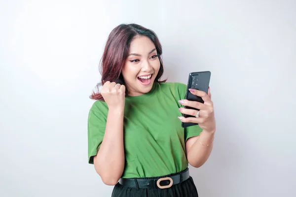 Mutlu yüzlü, yeşil tişört giyen ve elinde beyaz arkaplan ile izole akıllı telefon tutan Asyalı genç bir kadın.