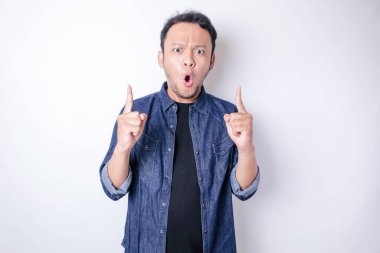 Şok olmuş Asyalı adam mavi donanma gömleği giyip beyaz arka planla izole edilmiş fotokopi alanını işaret ediyor.