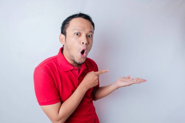 Kırmızı tişörtlü, şok olmuş Asyalı adam yanındaki fotokopi odasını işaret ediyor, beyaz bir arka planla izole edilmiş.