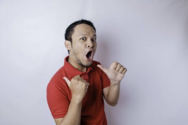 Kırmızı tişörtlü, şok olmuş Asyalı adam yanındaki fotokopi odasını işaret ediyor, beyaz bir arka planla izole edilmiş.