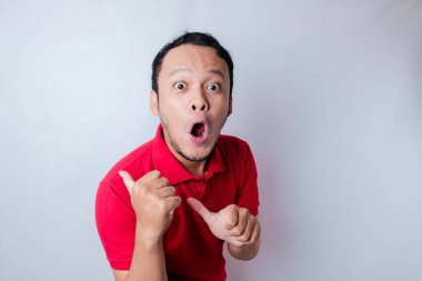 Kırmızı tişörtlü, şok olmuş Asyalı adam yanındaki fotokopi odasını işaret ediyor, beyaz bir arka planla izole edilmiş.
