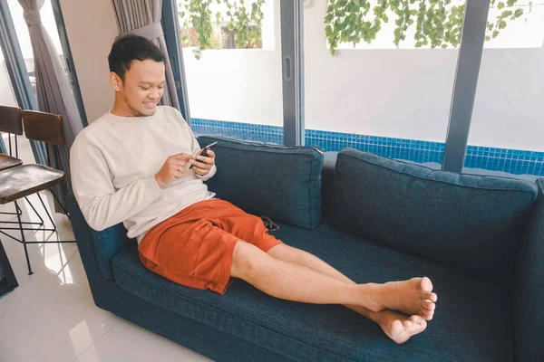 아시아인 남자는 집에서 소파에 앉아서 스마트폰으로 휴대폰 메시지를 보내며 시간을 — 스톡 사진
