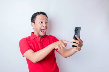 Kırmızı tişörtlü, beyaz arka planla izole edilmiş akıllı telefonunu işaret eden Asyalı bir adam.