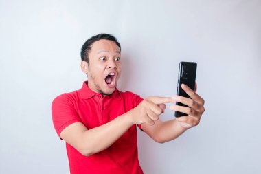 Kırmızı tişörtlü, beyaz arka planla izole edilmiş akıllı telefonunu işaret eden Asyalı bir adam.