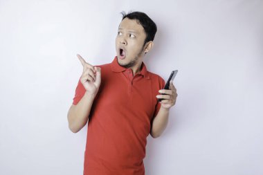 Kırmızı tişörtlü, beyaz arka planla izole edilmiş akıllı telefonunu işaret eden Asyalı bir adam.