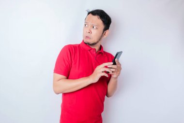 Kırmızı tişörtlü, beyaz arka planla izole edilmiş akıllı telefonunu işaret eden Asyalı bir adam.
