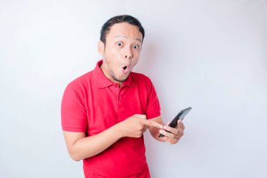 Kırmızı tişörtlü, beyaz arka planla izole edilmiş akıllı telefonunu işaret eden Asyalı bir adam.