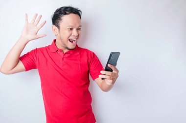 Kırmızı tişörtlü, beyaz arka planla izole edilmiş akıllı telefonunu işaret eden Asyalı bir adam.