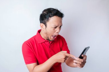Kızgın genç Asyalı bir adam, elinde telefonuyla kırmızı tişört giymiş sinirli bir yüz ifadesi takınıyor.