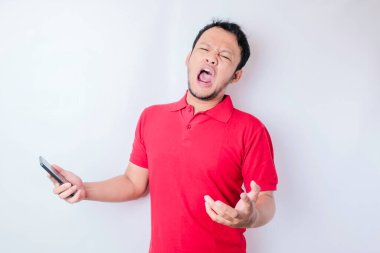 Kızgın genç Asyalı bir adam, elinde telefonuyla kırmızı tişört giymiş sinirli bir yüz ifadesi takınıyor.