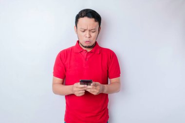 Memnuniyetsiz bir Asyalı adam, elinde telefonuyla kırmızı tişört giymiş, tedirgin bir yüz ifadesi takınıyor.