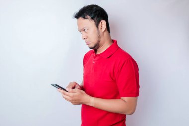 Memnuniyetsiz bir Asyalı adam, elinde telefonuyla kırmızı tişört giymiş, tedirgin bir yüz ifadesi takınıyor.