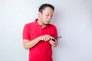 Memnuniyetsiz bir Asyalı adam, elinde telefonuyla kırmızı tişört giymiş, tedirgin bir yüz ifadesi takınıyor.
