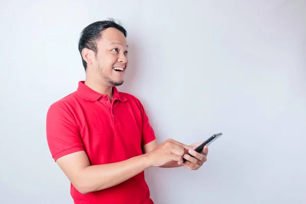 白い背景によって隔離された 彼のスマートフォンを指して赤いTシャツを身に着けている驚きのアジアの男 — ストック写真