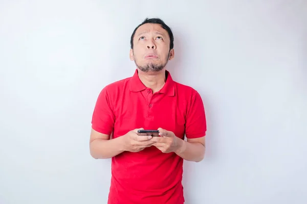 不満な若いアジアの男は彼の携帯電話を保持赤いTシャツのいらいら顔の表情を着て不満そうに見えます — ストック写真