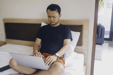 Yorgun genç adam yatağında otururken laptopunu kontrol ediyor.