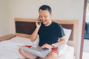 Yatakta çalışan mutlu genç adam. Dizlerinin üzerine çök ve telefonda konuş..