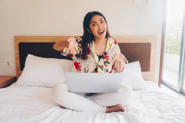 Yataktan çalışan mutlu genç bir kadın. Dizlerinin üzerine çök ve klavyeye yaz..