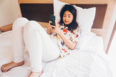 Uykulu genç bir kadın telefonu yatakta tutarken esniyor..